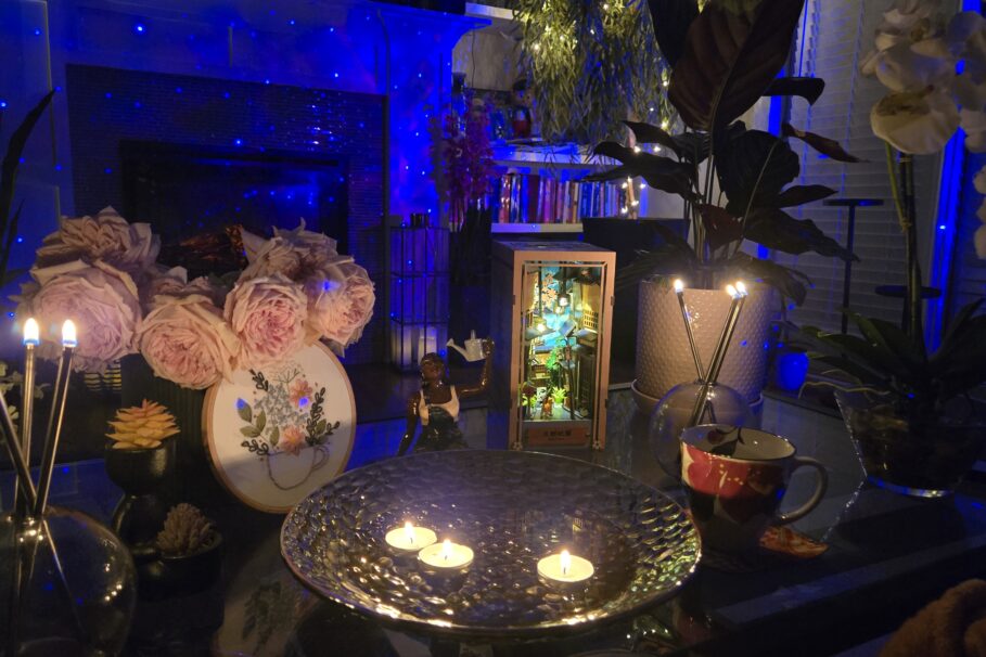 A cozy, dimly lit interior vignette bathed in deep blue ambient light from a star projector or string lights in the background. A glass coffee table holds a decorative silver hammered bowl with three lit tea light candles at its center. Surrounding it: a bouquet of full, blush pink garden roses; an embroidery hoop displaying a floral teacup design in progress; a small dark figurine of a seated woman; a lit book nook diorama in a wooden frame depicting a detailed miniature alleyway scene; a textured white ceramic vase holding tall taper candles; a decorative mug with floral motif; and various plants including tropical leaves and white orchids. In the background, a bookshelf glows with warm string lights, and trailing greenery hangs from above. The overall atmosphere is intimate and curated.